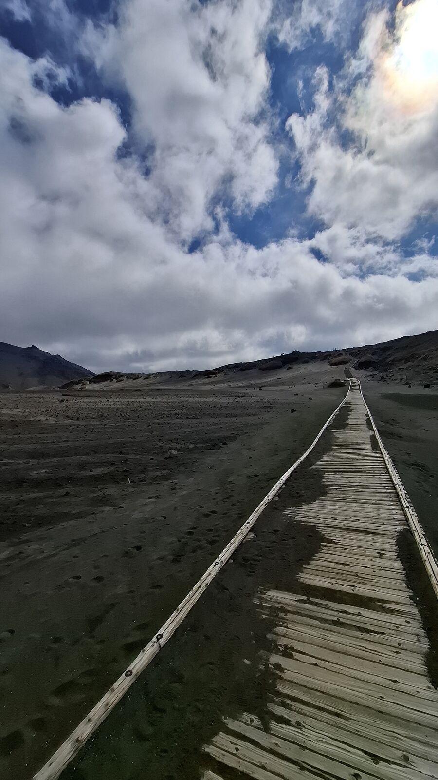 Long boardwalk stretching across the volcanic desert, no vegetation, just grey ash and rock and sky, the path leading toward the distant caldera wall