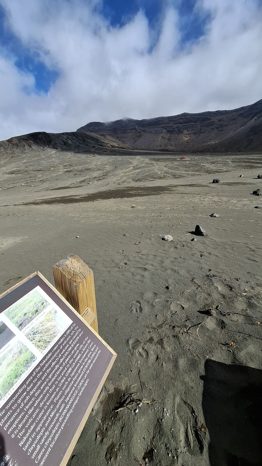 Information board standing in the volcanic desert, black ash and dark crater walls behind it, explaining the geology of the area