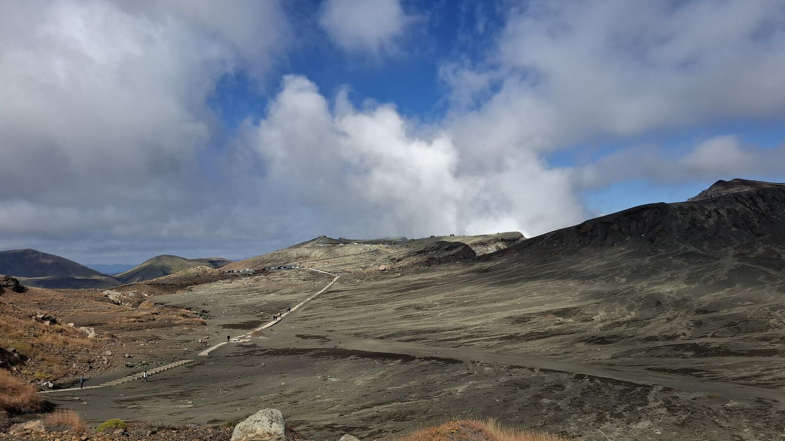 Wide panorama of the volcanic desert with a boardwalk path leading across grey ash toward the steaming crater in the distance, clouds and blue sky above