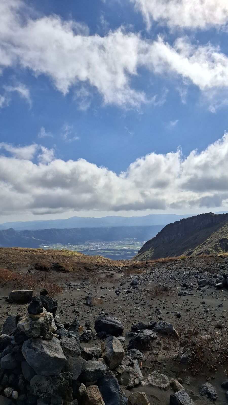Rocky volcanic terrain with scattered boulders, the caldera rim visible in the distance, clouds casting shadows across the barren ground