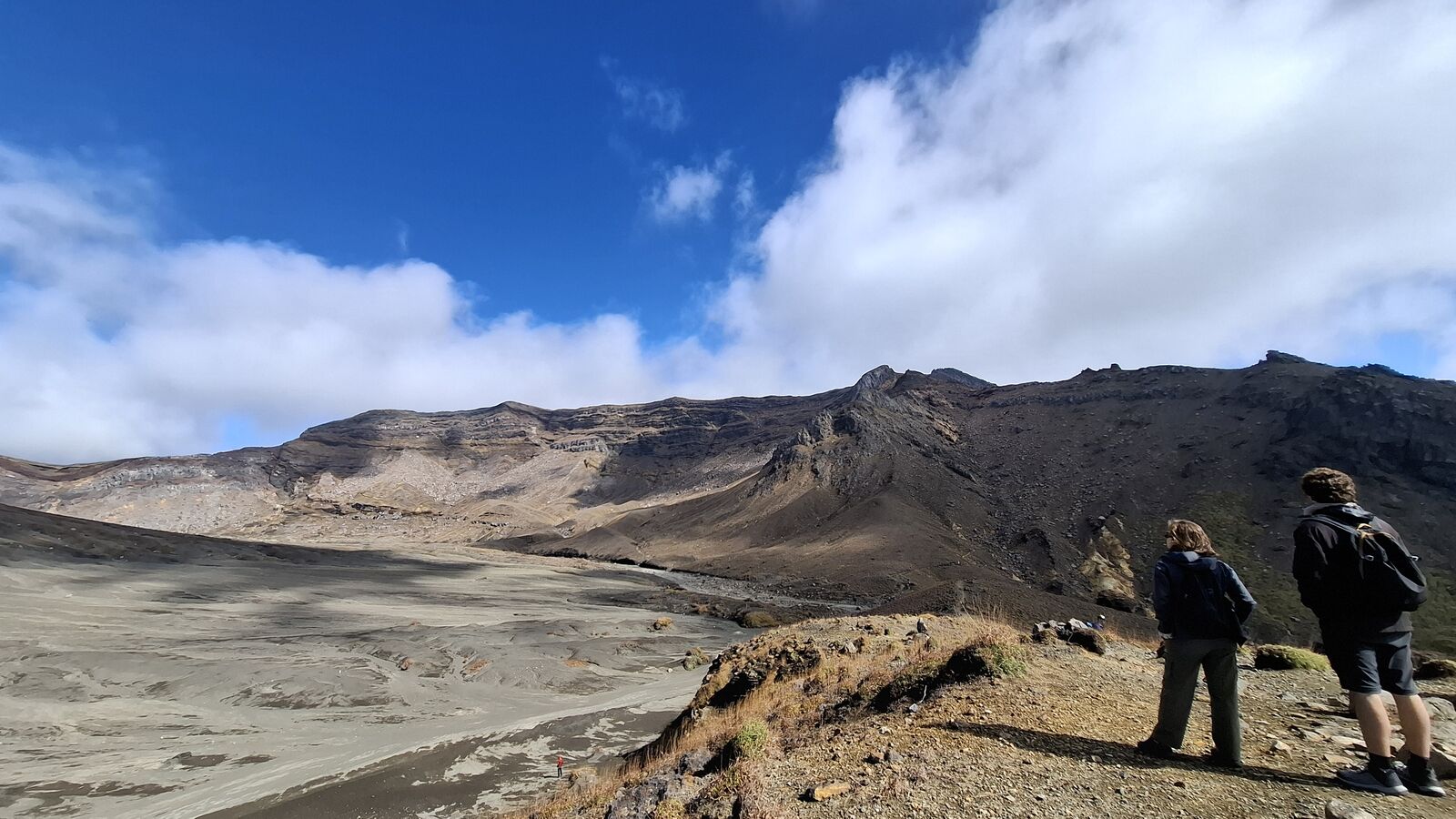 Two hikers standing at the edge of the caldera looking out over the vast volcanic plain, the scale of the landscape visible in their small silhouettes