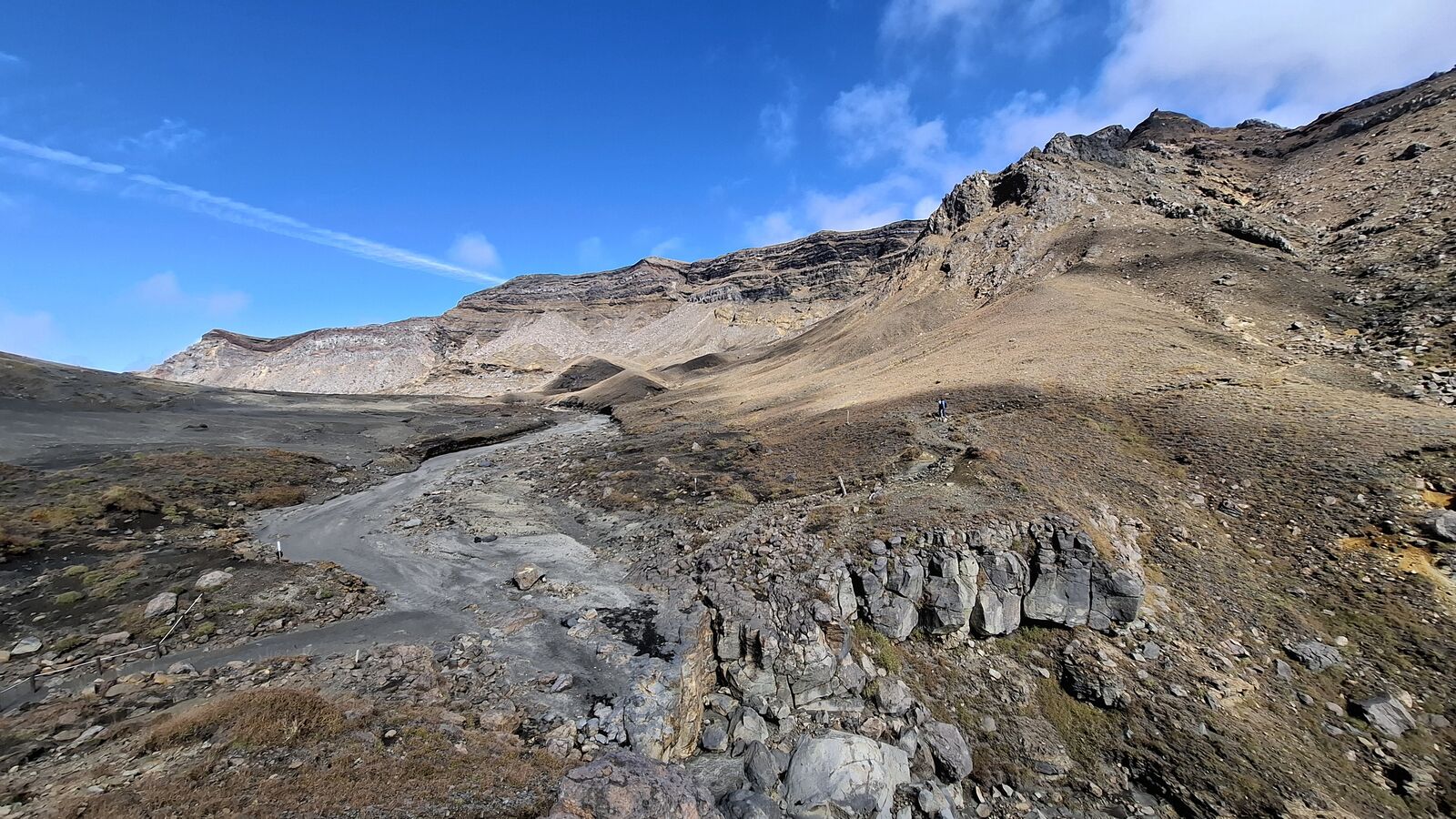 The caldera floor with layered volcanic rock, dried stream beds cutting through the ash, blue sky and distant peaks framing the scene