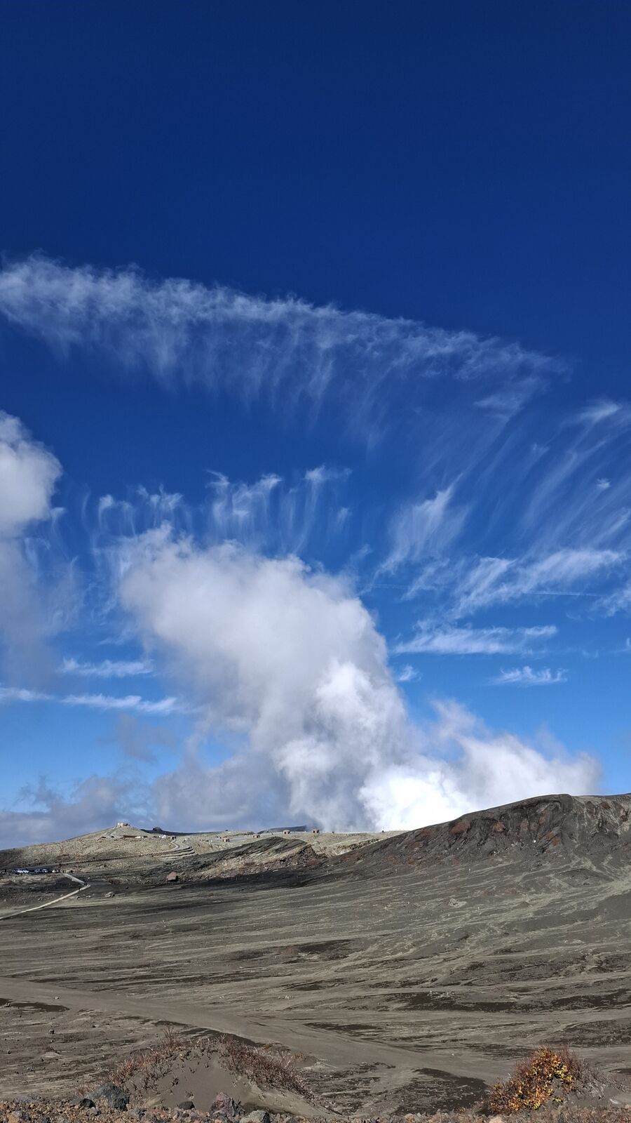 Massive steam cloud rising from Mount Aso's crater, tiny bunkers on the ridgeline showing the enormous scale of Japan's largest active volcano