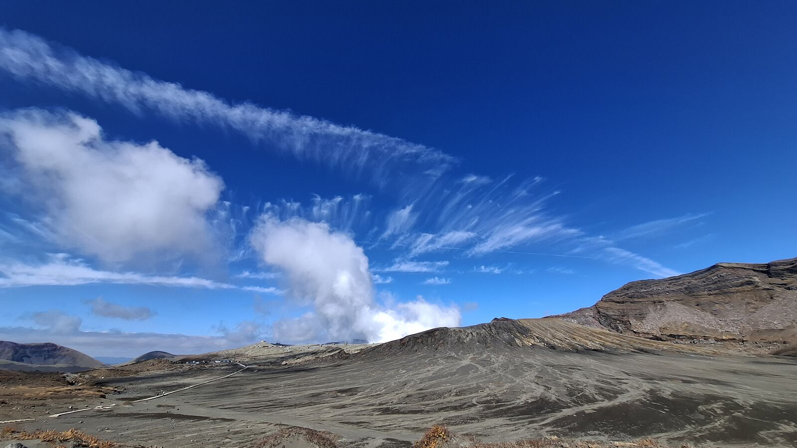 Steam plume rising from Mount Aso's Nakadake crater against a deep blue sky with cirrus clouds, barren volcanic landscape stretching to the horizon