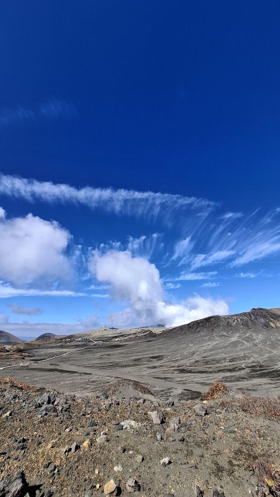 The volcanic landscape from higher ground, steam column and barren ridgeline stretching across the frame, cirrus clouds streaming overhead