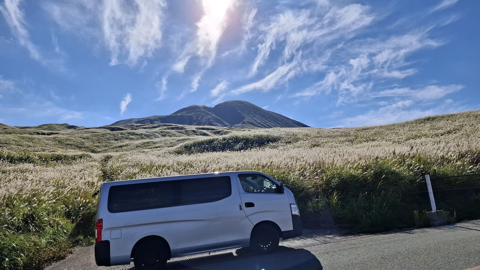The camper van parked at the edge of golden susuki grass fields, the volcano cone rising perfectly behind it, a sea of silver-gold grass swaying in the wind under dramatic clouds