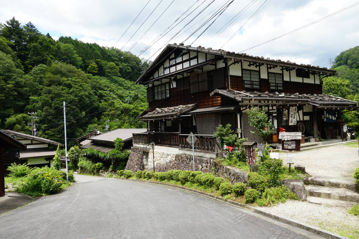 A traditional timber-framed house with steep tiled roof and dark wood construction, nestled among green rice paddies in the Kiso Valley
