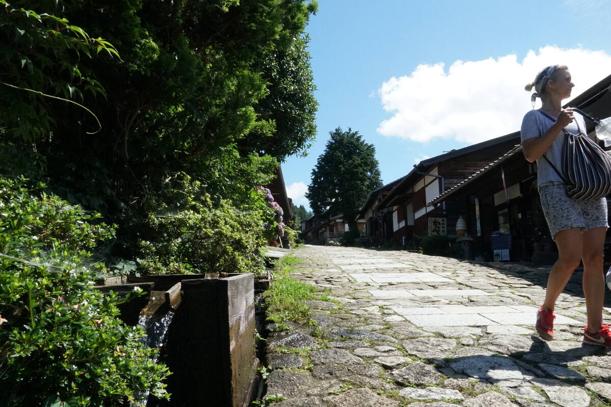 A hiker pauses on the stone-paved path through Tsumago with traditional shops and dark timber buildings visible