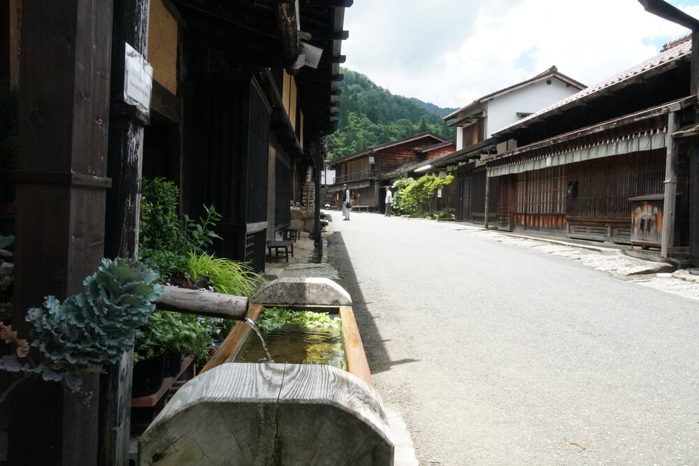 Wooden water trough with plants in front of traditional buildings, mountains in the background