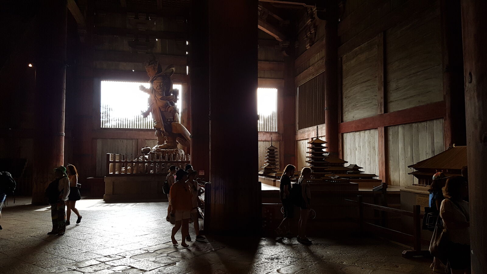 The dark, atmospheric interior of Todai-ji, silhouettes of visitors against bright window openings, a guardian figure catching the light, the vast space swallowing sound and scale