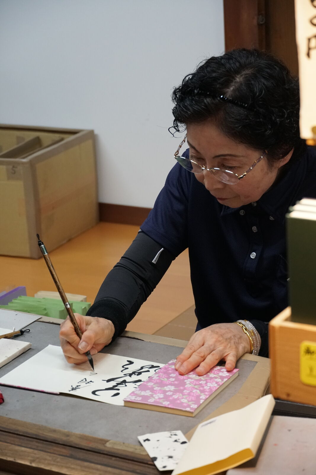 A woman in dark robes carefully painting a goshuin temple stamp book with calligraphy brush, concentration and skill in every stroke