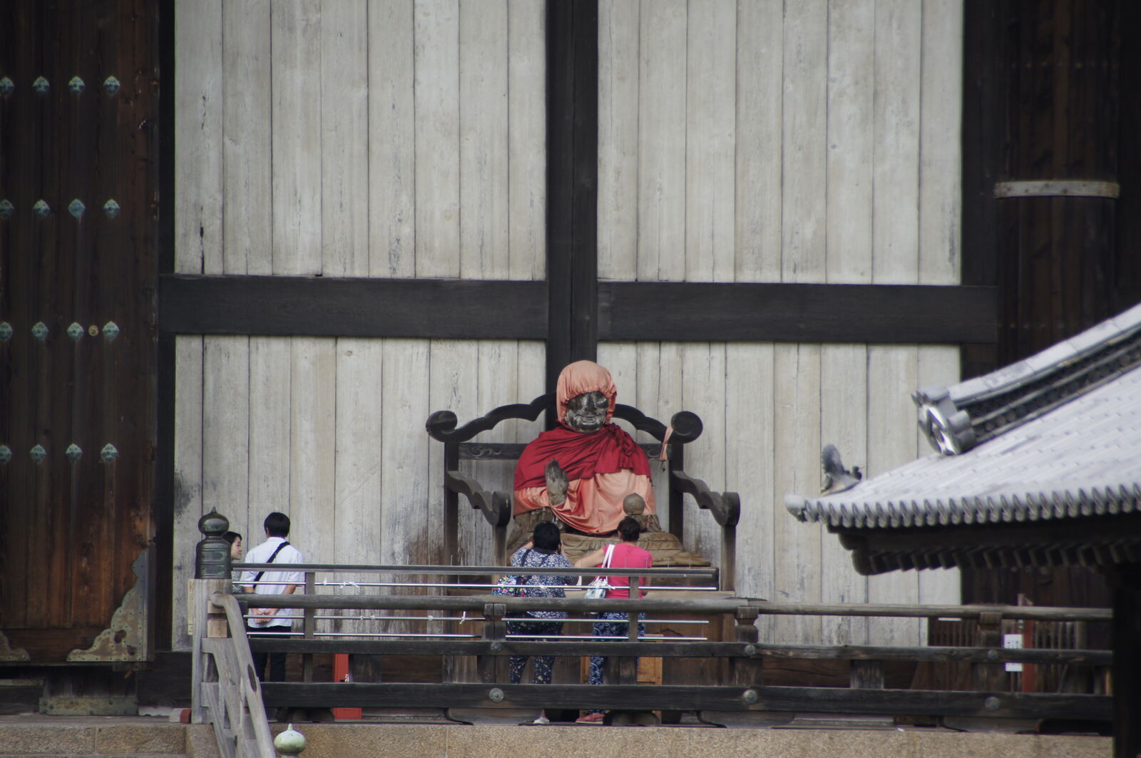 A seated figure in red robes on a wooden platform against the weathered facade of the Daibutsuden, the ancient wood panels stretching up behind