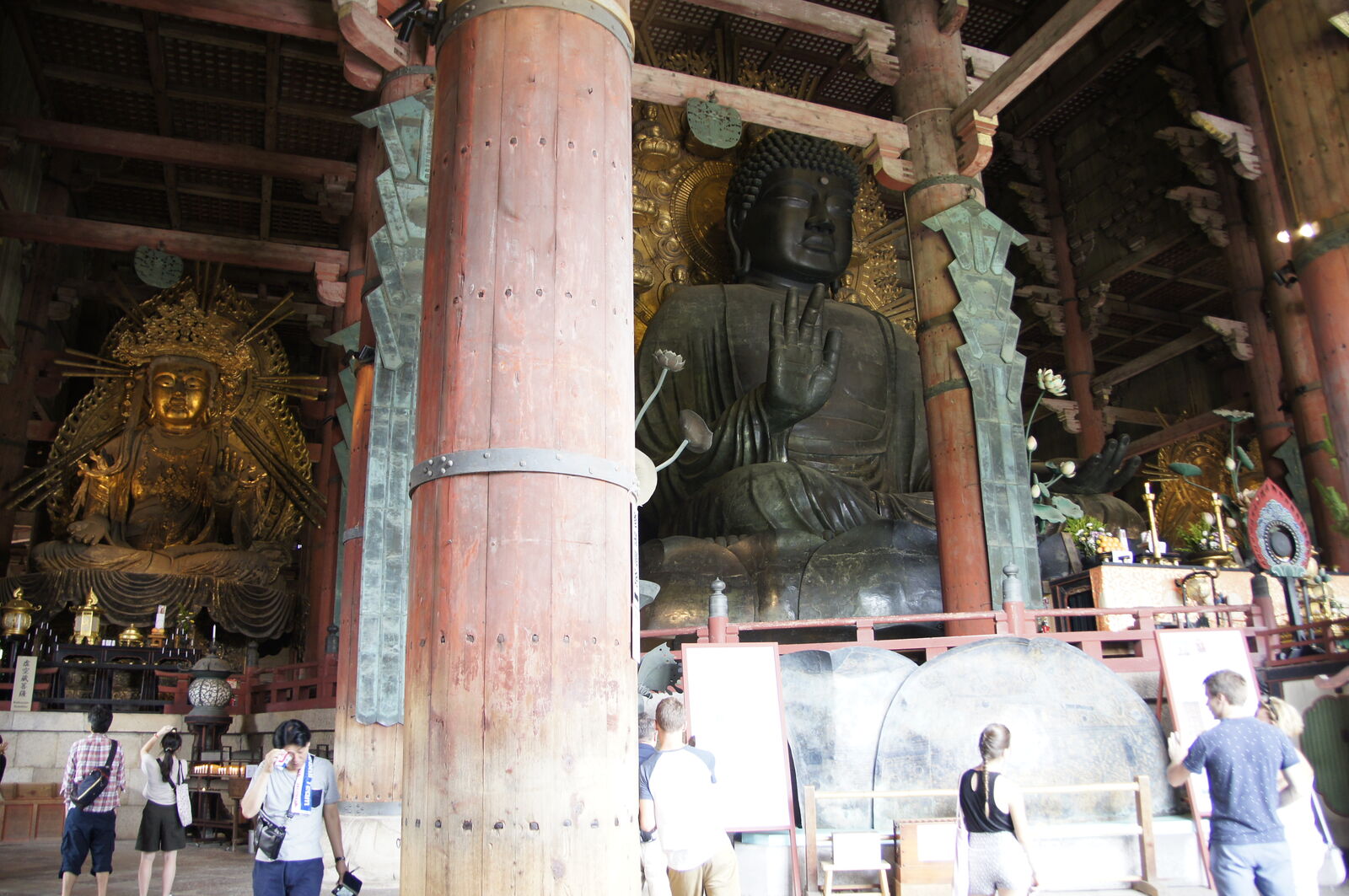 Inside the Daibutsuden: the Great Buddha flanked by a golden Kannon figure, visitors looking up in awe, the red-painted wooden pillars reaching into the shadowy ceiling above