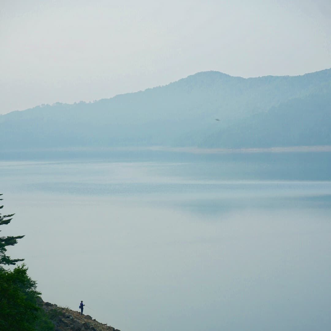 Lone fisherman at Lake Nozori in the early morning mist, mountains fading into haze across perfectly still water