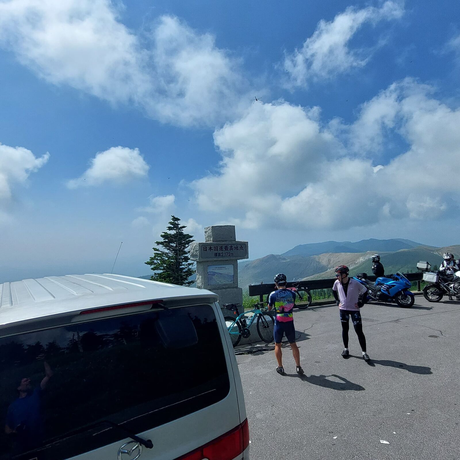 Busy scene at the 2,172m monument: campervan, cyclists resting, motorcycles parked, and mountain ridges stretching into the distance
