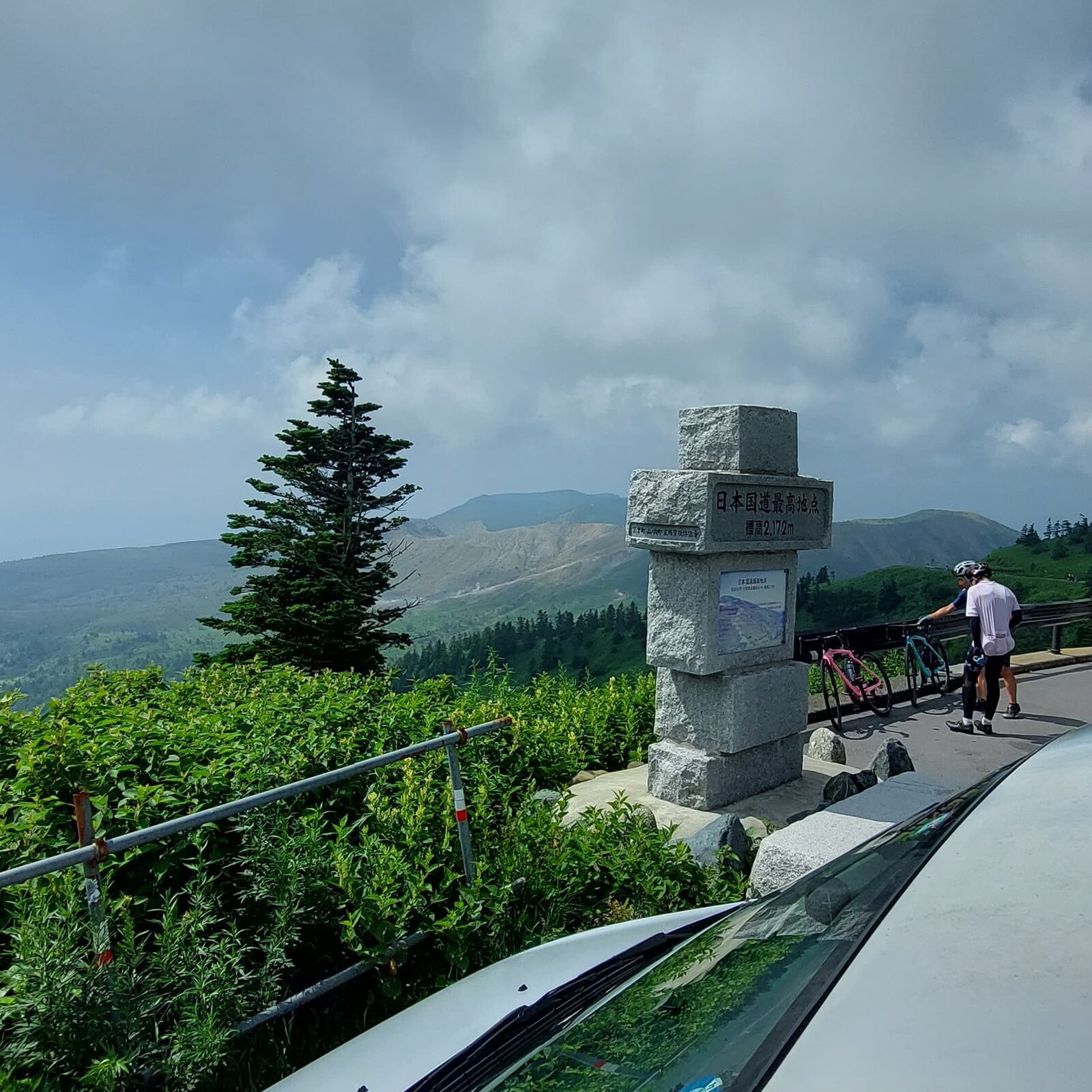 The campervan at the monument with a lone cyclist in the background and clouds rolling over the peaks