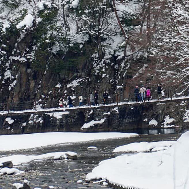 People crossing a suspension bridge over the river with snow-covered cliffs and forest on both sides