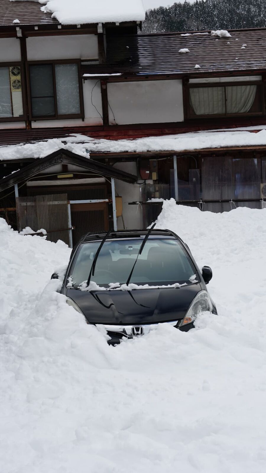 A car almost completely buried in snow beside a traditional house, showing the depth of winter accumulation
