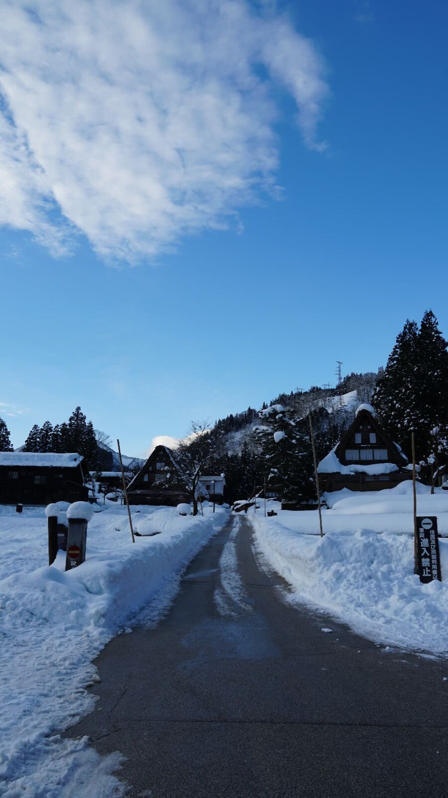 Village entrance on a clear winter morning, blue sky above, gassho rooftops heavy with snow, mountains in the distance