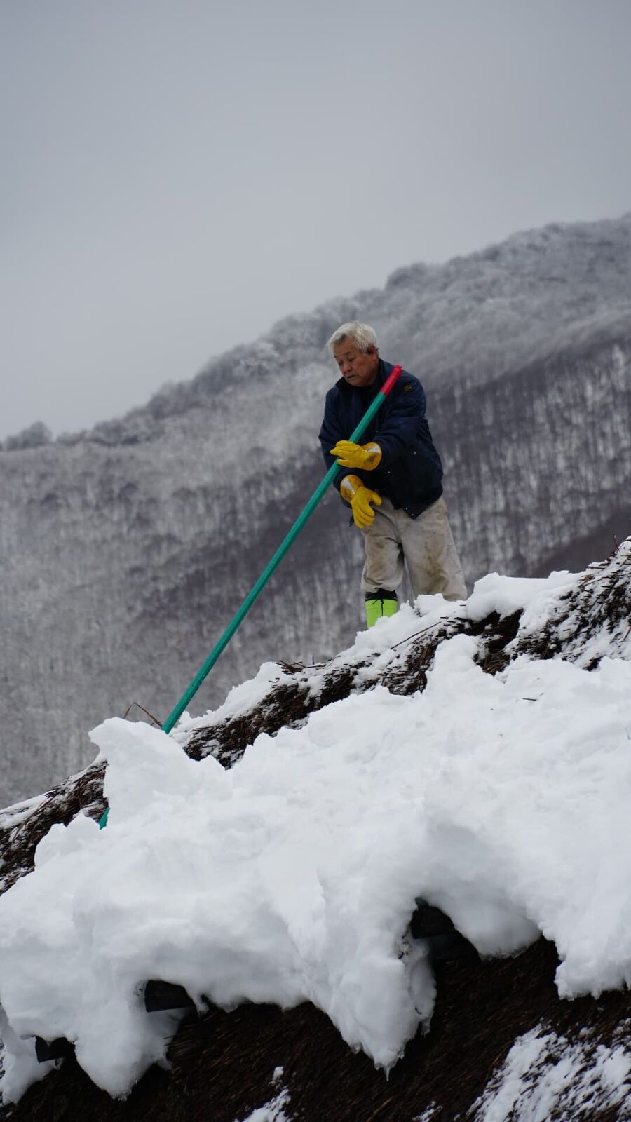 Worker on a traditional house roof pushing snow off the edge, cascade of white falling below