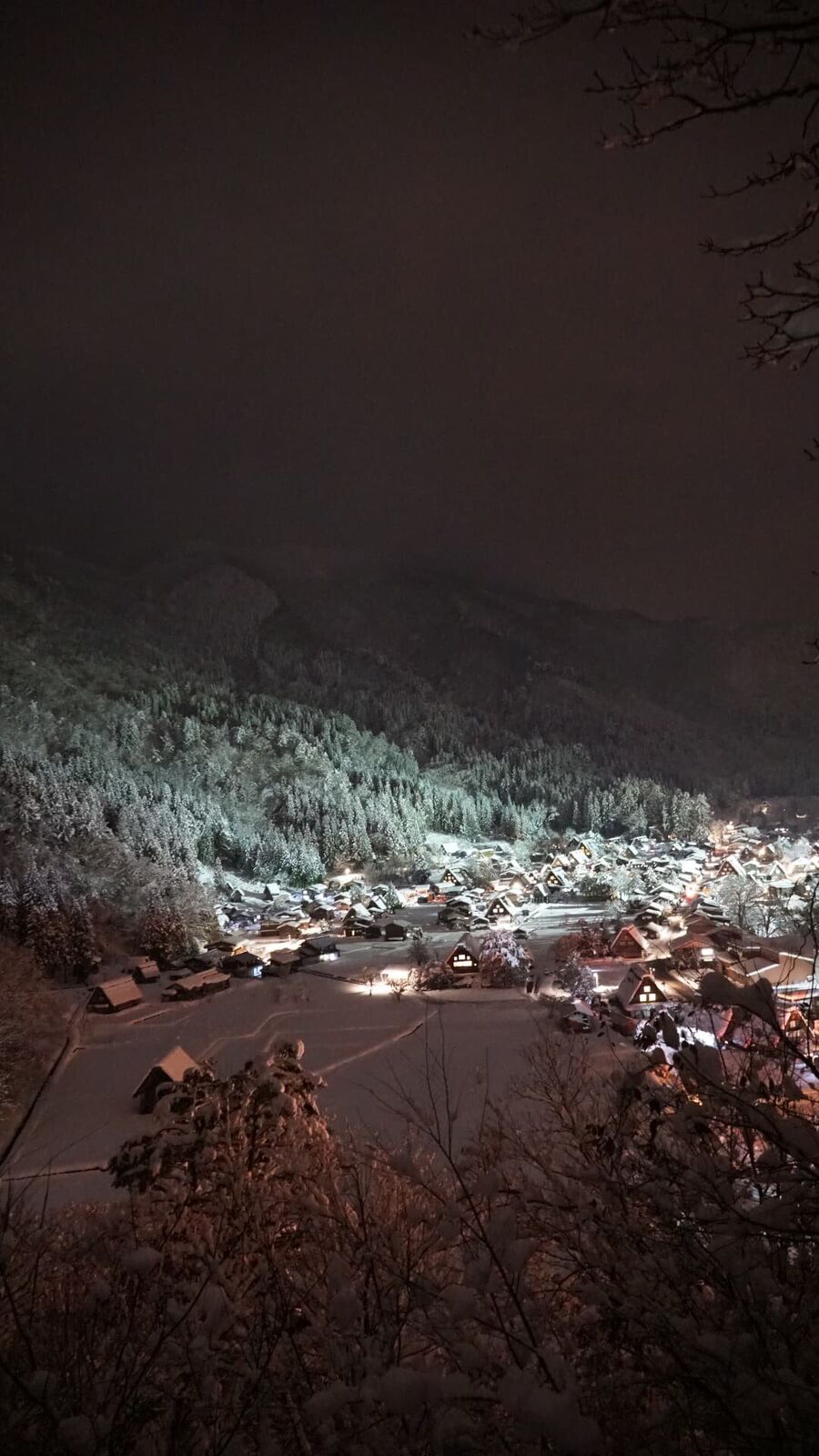Wider night illumination panorama from the viewpoint, the entire village lit up against snow-covered forest and dark sky