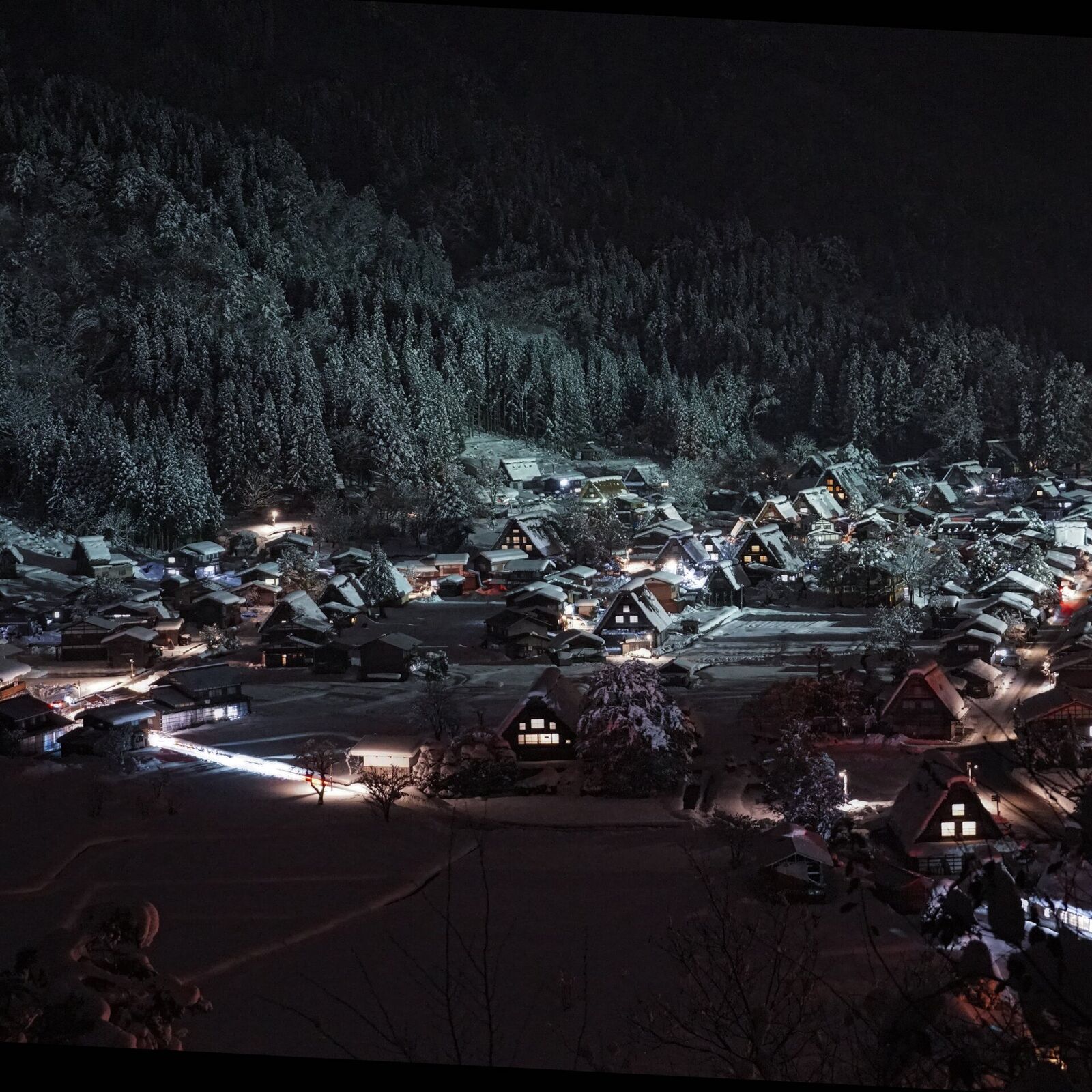 Tighter view of the illuminated village at night, individual Gassho roofs and warm window lights clearly visible against the snow
