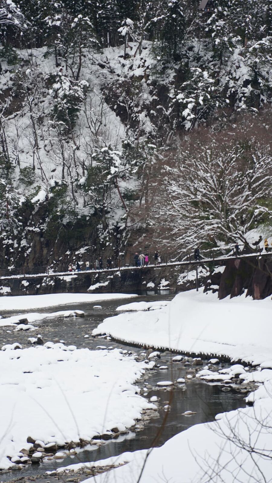 Visitors crossing a narrow bridge over the river, snow-laden forest and mountains forming the backdrop