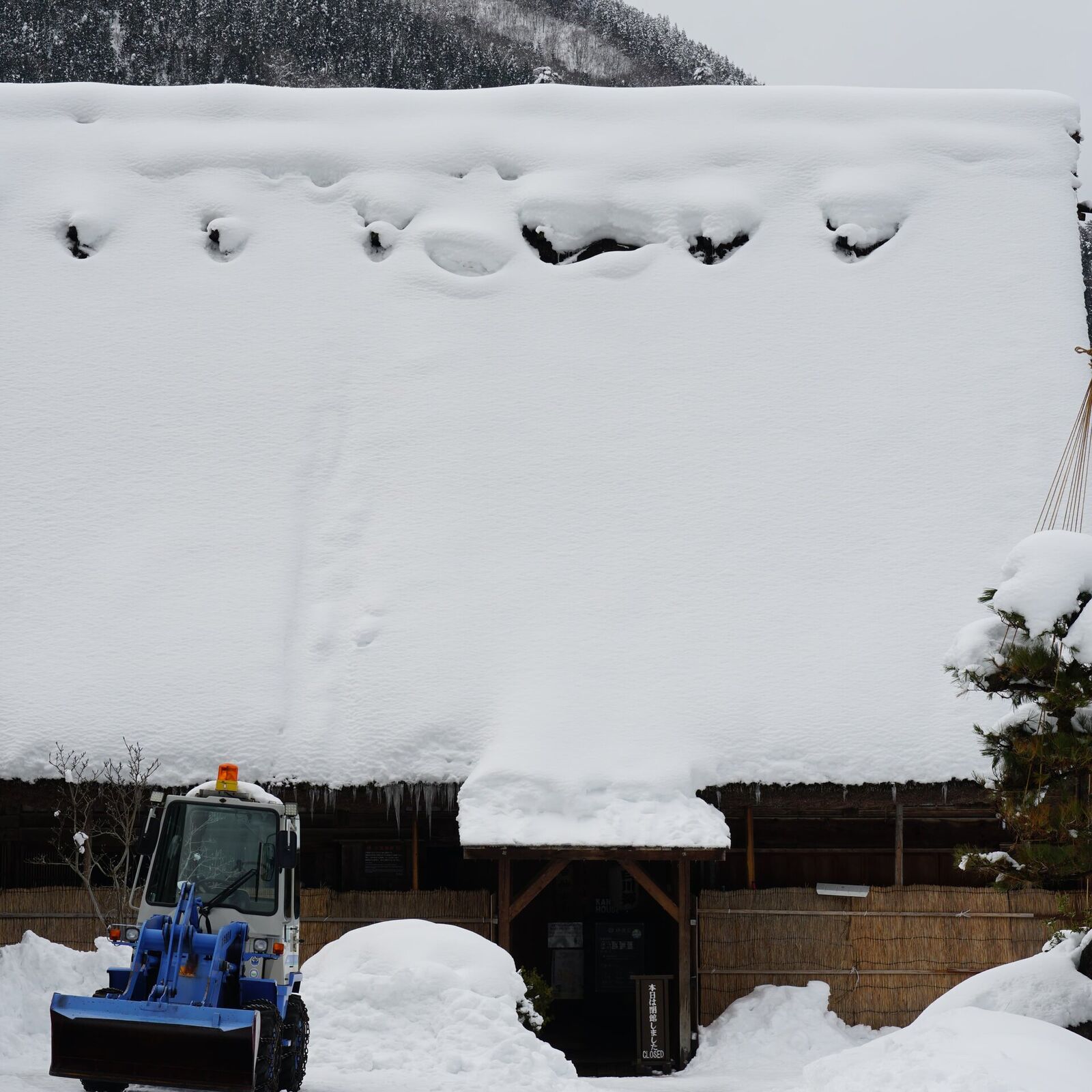 Elderly man balanced on a steep Gassho-zukuri roof clearing heavy snow with a long tool, mountains behind