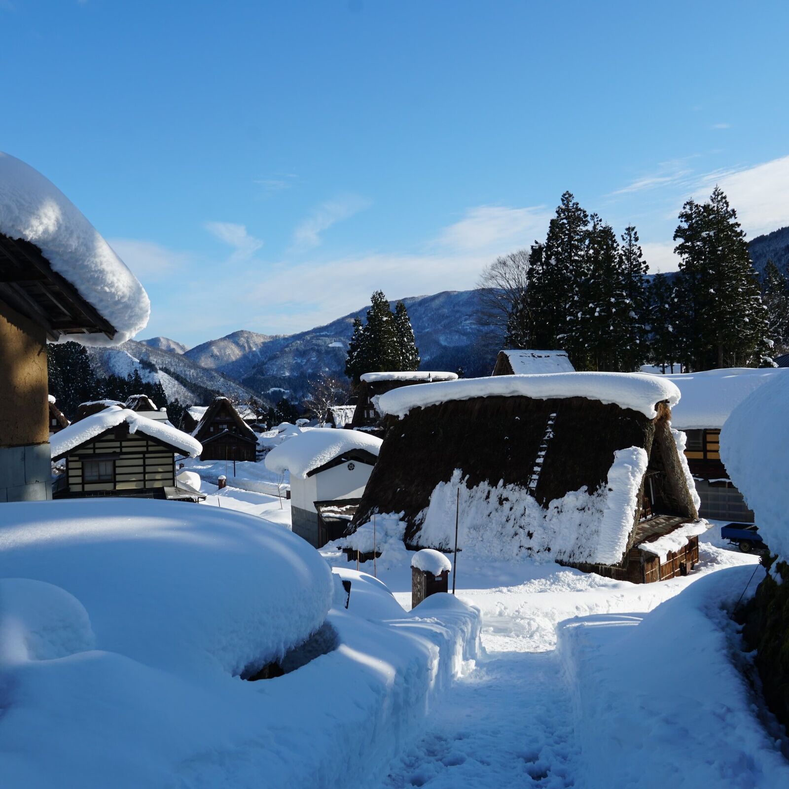 Village rooftops buried under meters of snow on a clear blue-sky morning, rounded snow formations and dark thatched roofs