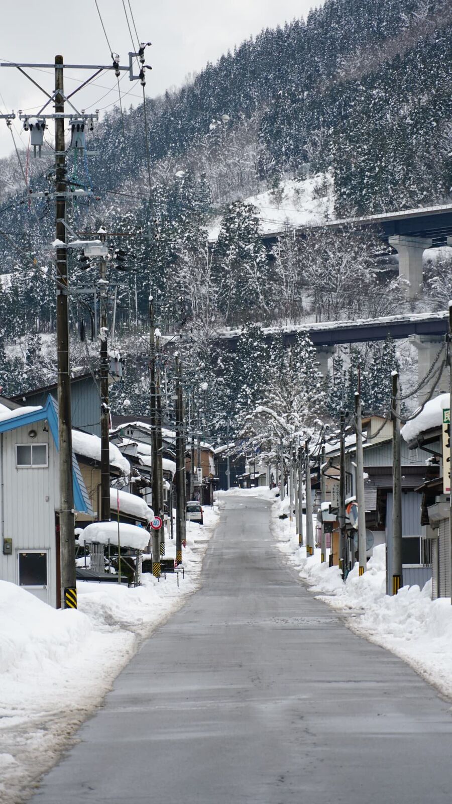 Snowy village street stretching into the distance, buildings buried in white, mountains and falling snow behind