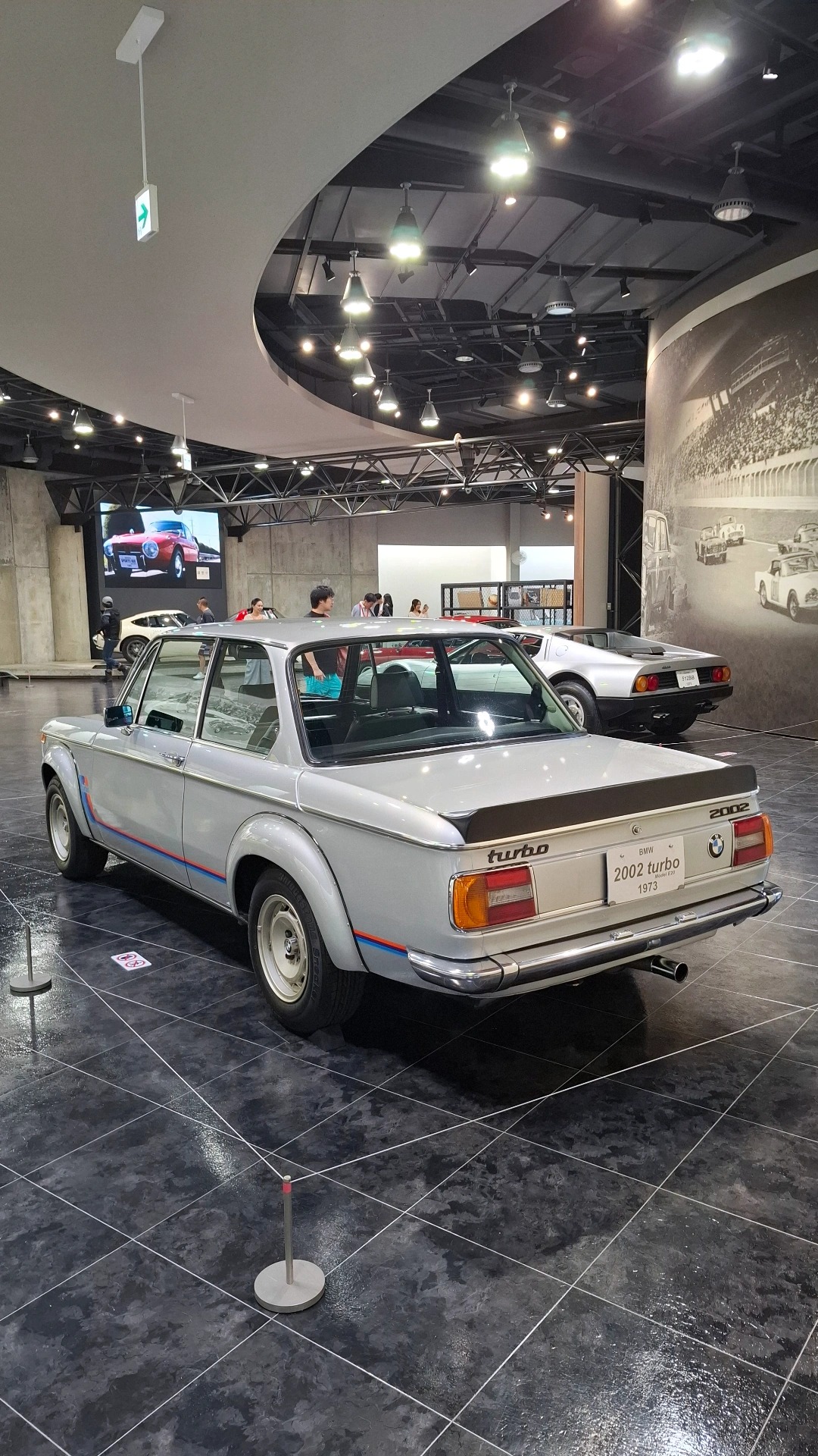 1973 BMW 2002 Turbo rear three-quarter view, white with M-stripes, Europe's first turbo production car, Audi Quattro visible behind