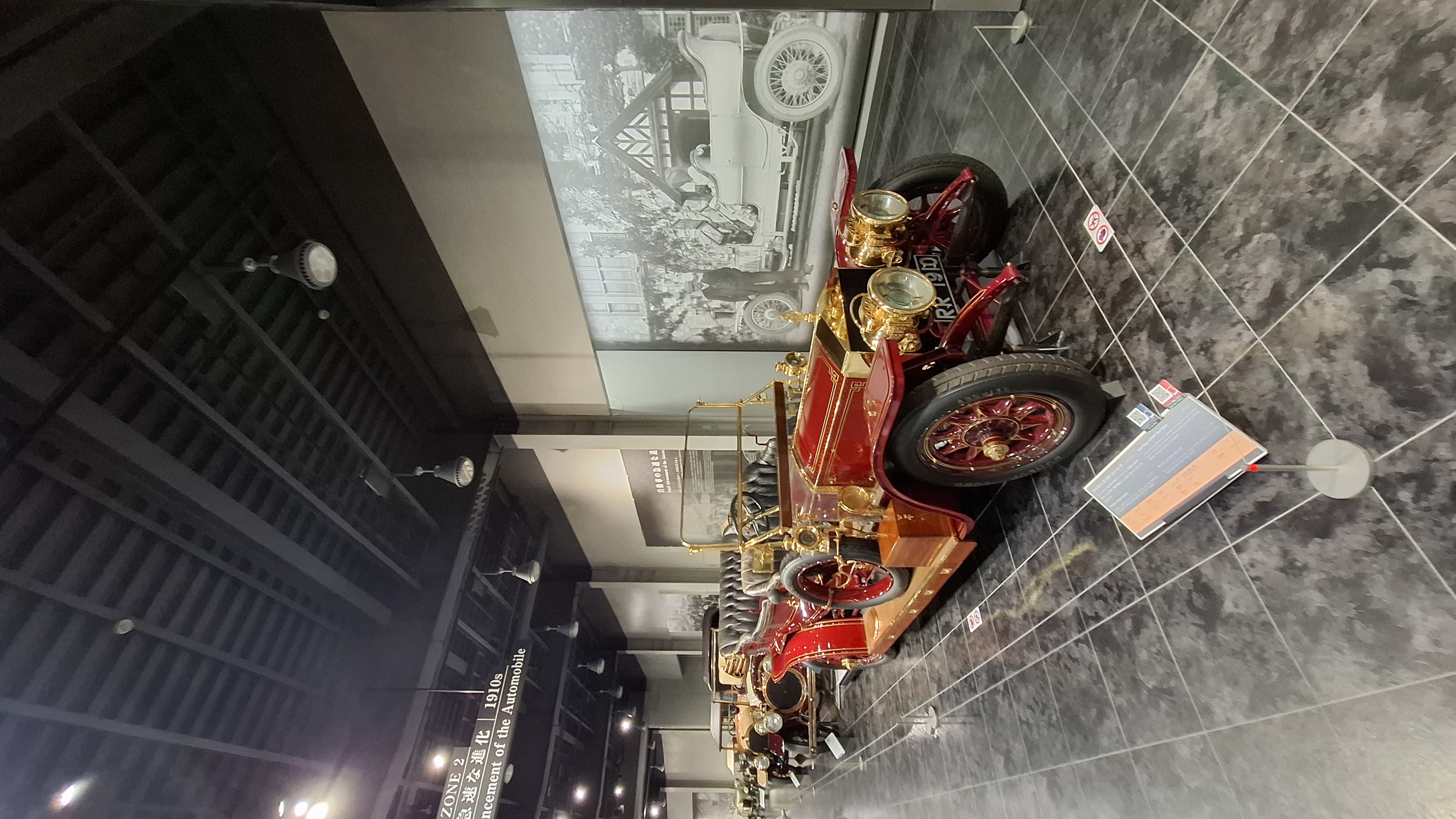 Red brass-era automobile with gold fittings and wooden wheels, period photographs on the wall behind