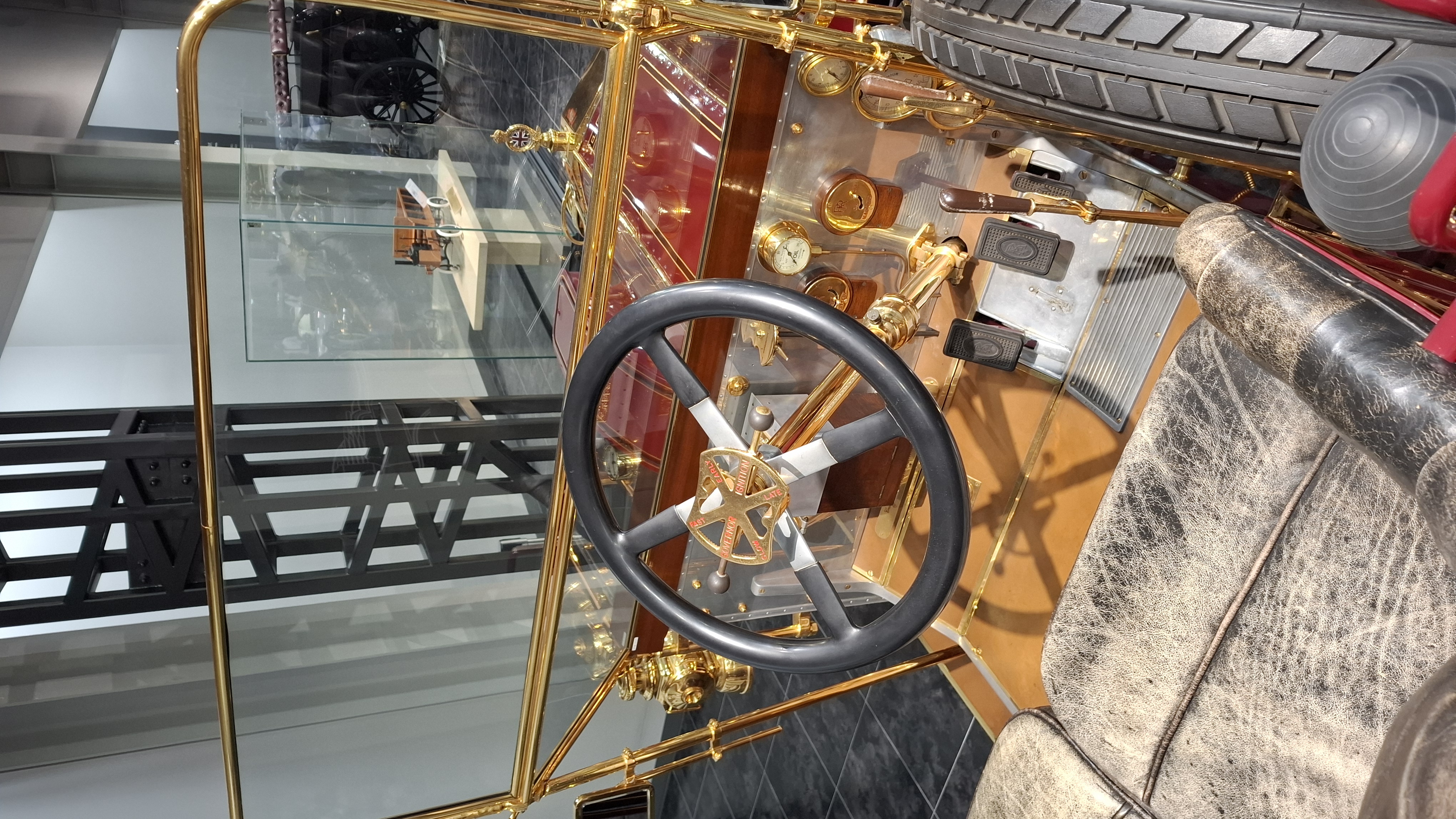 Brass-era car cockpit detail: tiller steering wheel, brass instruments, and leather seats