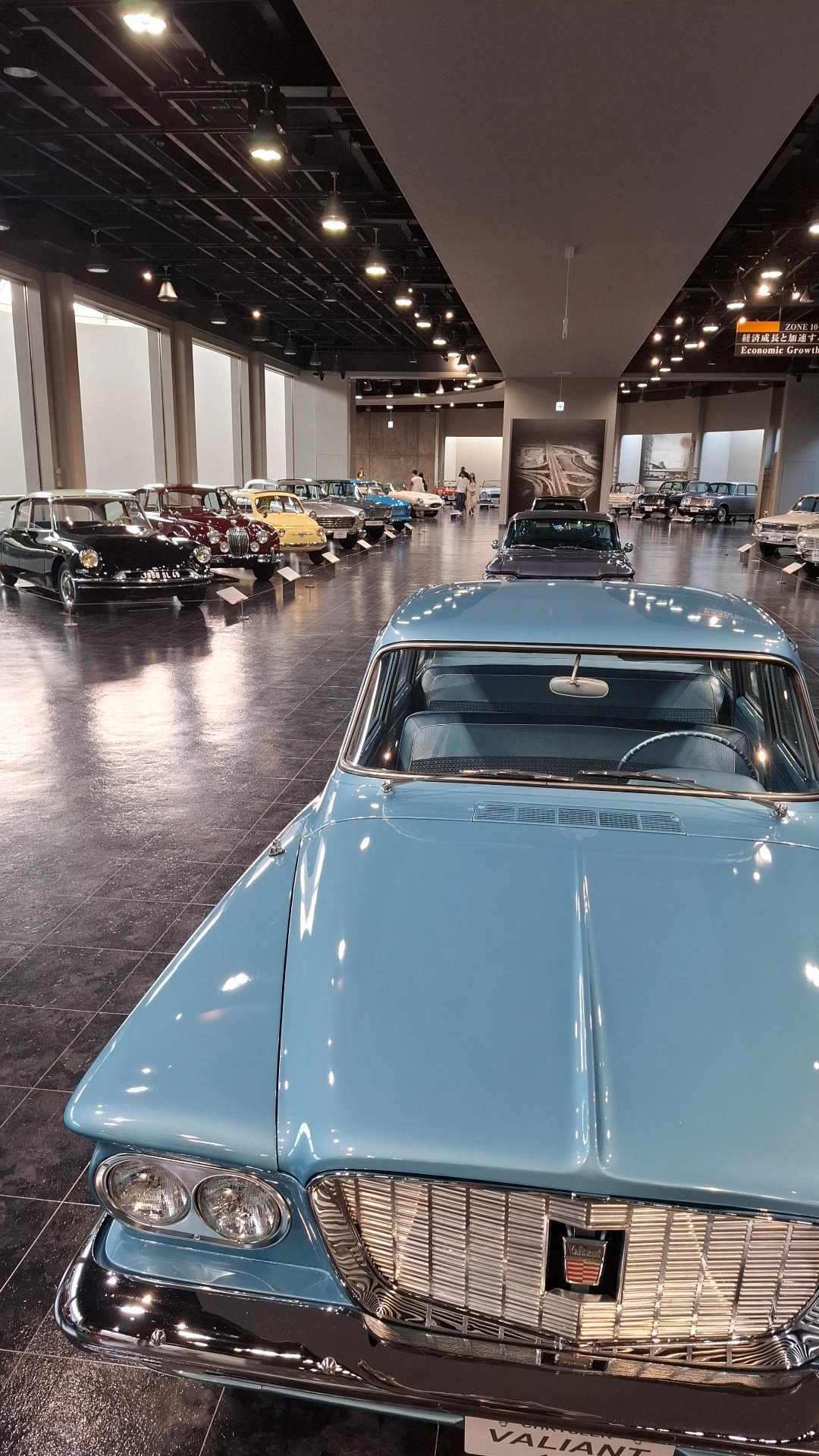 Light blue Plymouth Valiant leading a long row of classic cars stretching into the distance on polished dark tile