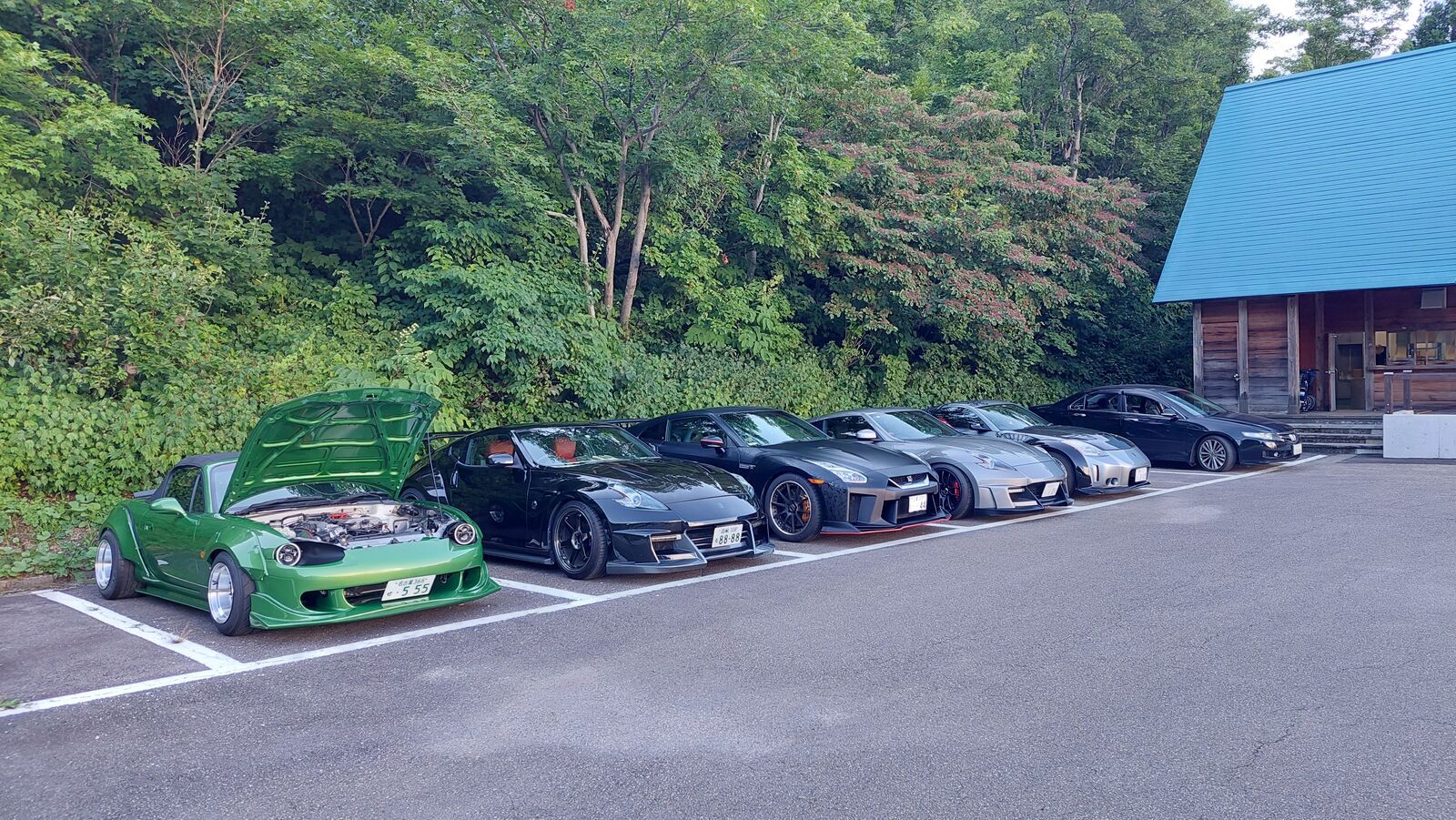 Row of modified Japanese sports cars parked at the summit: green Mazda MX-5 with hood up, Nissan 350Zs, and a GT-R