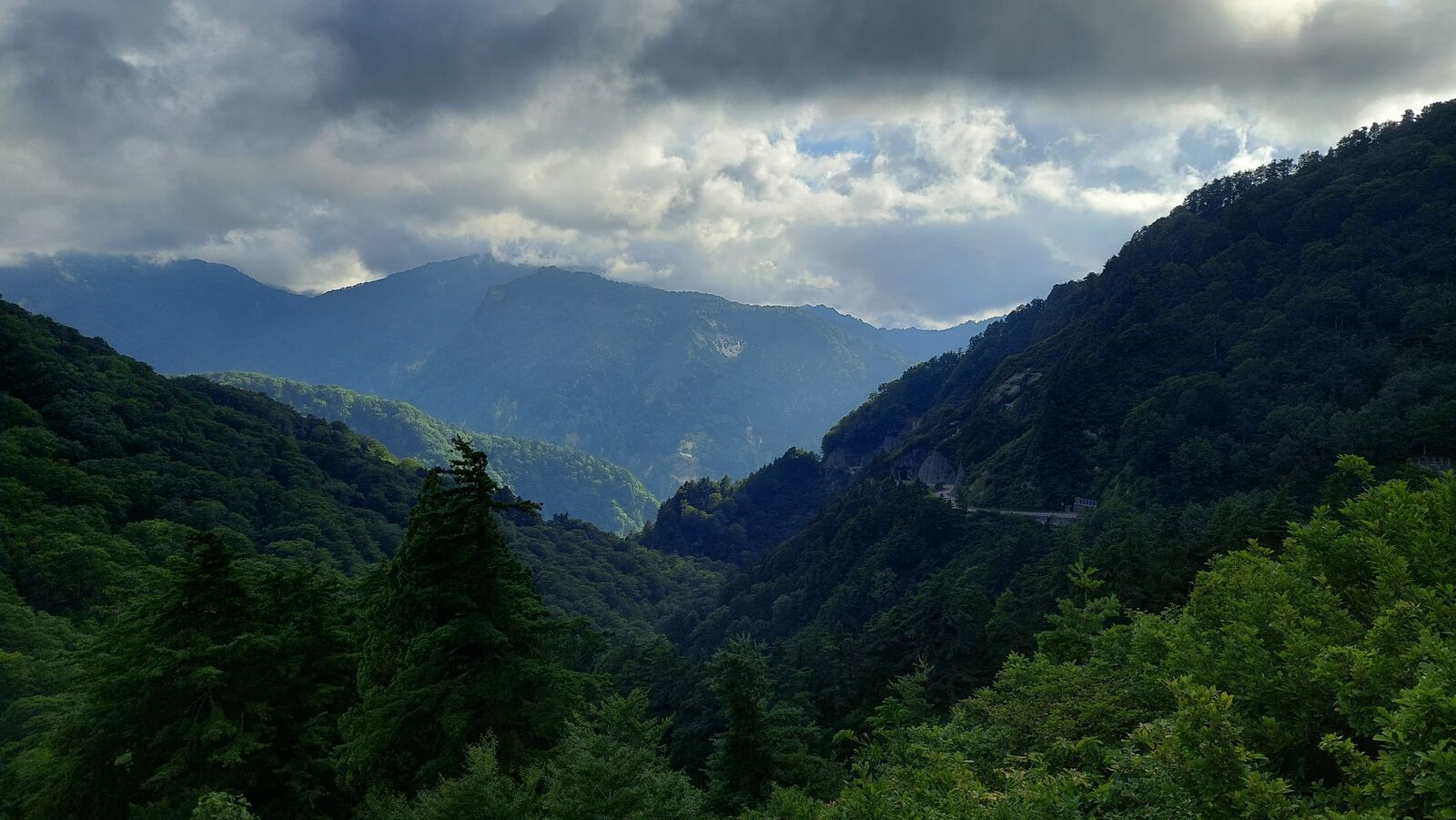 Dramatic mountain panorama with layered ridges, moody clouds, and the road carved into the far slope visible in the distance