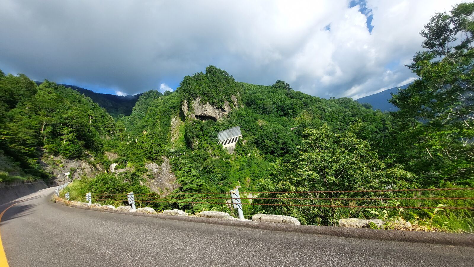 The road curving past exposed rock and built-in protective structures, green mountains rising behind