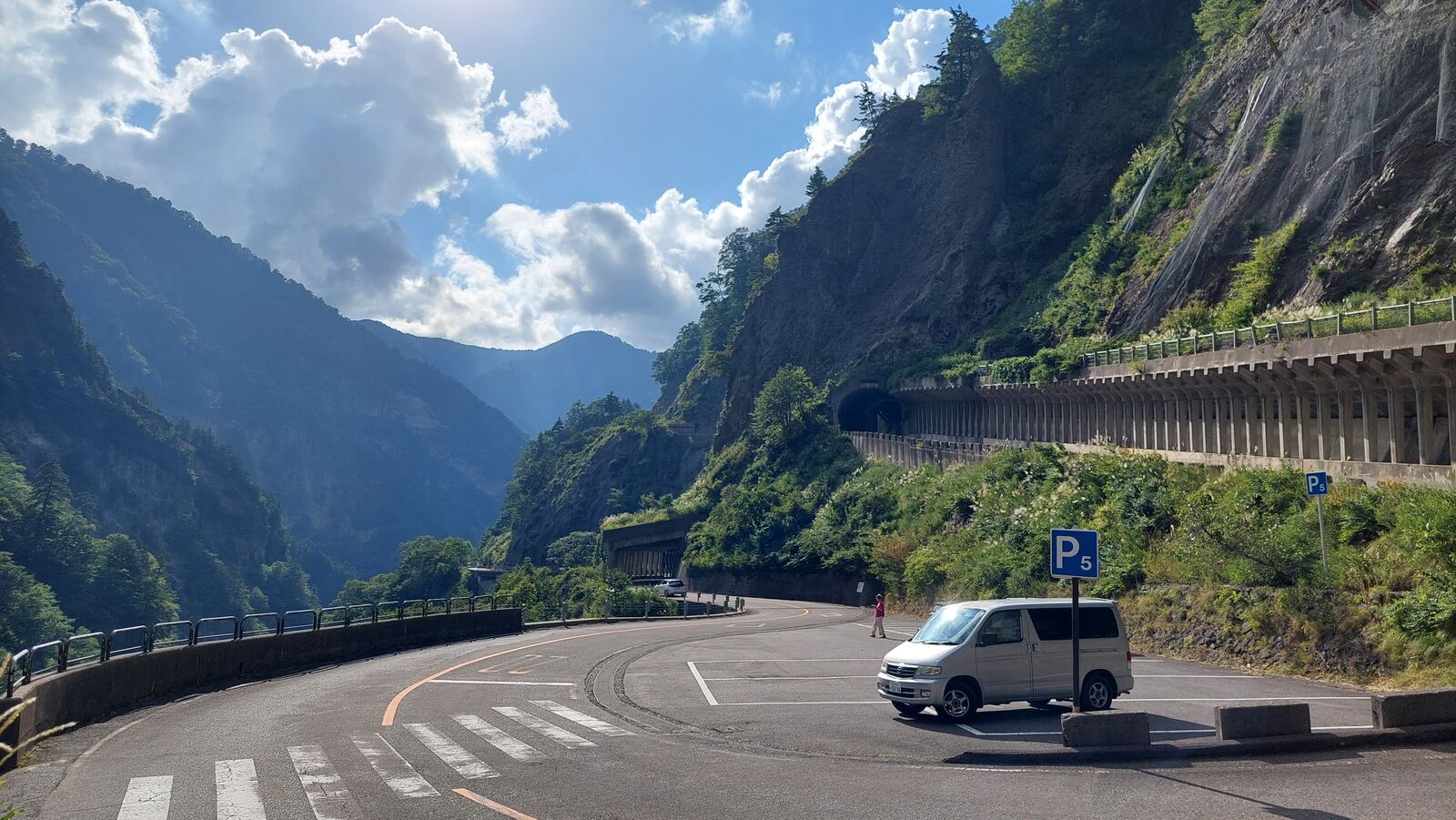 Campervan parked in a sweeping curve with a tunnel entrance ahead and mountain peaks framed by dramatic clouds